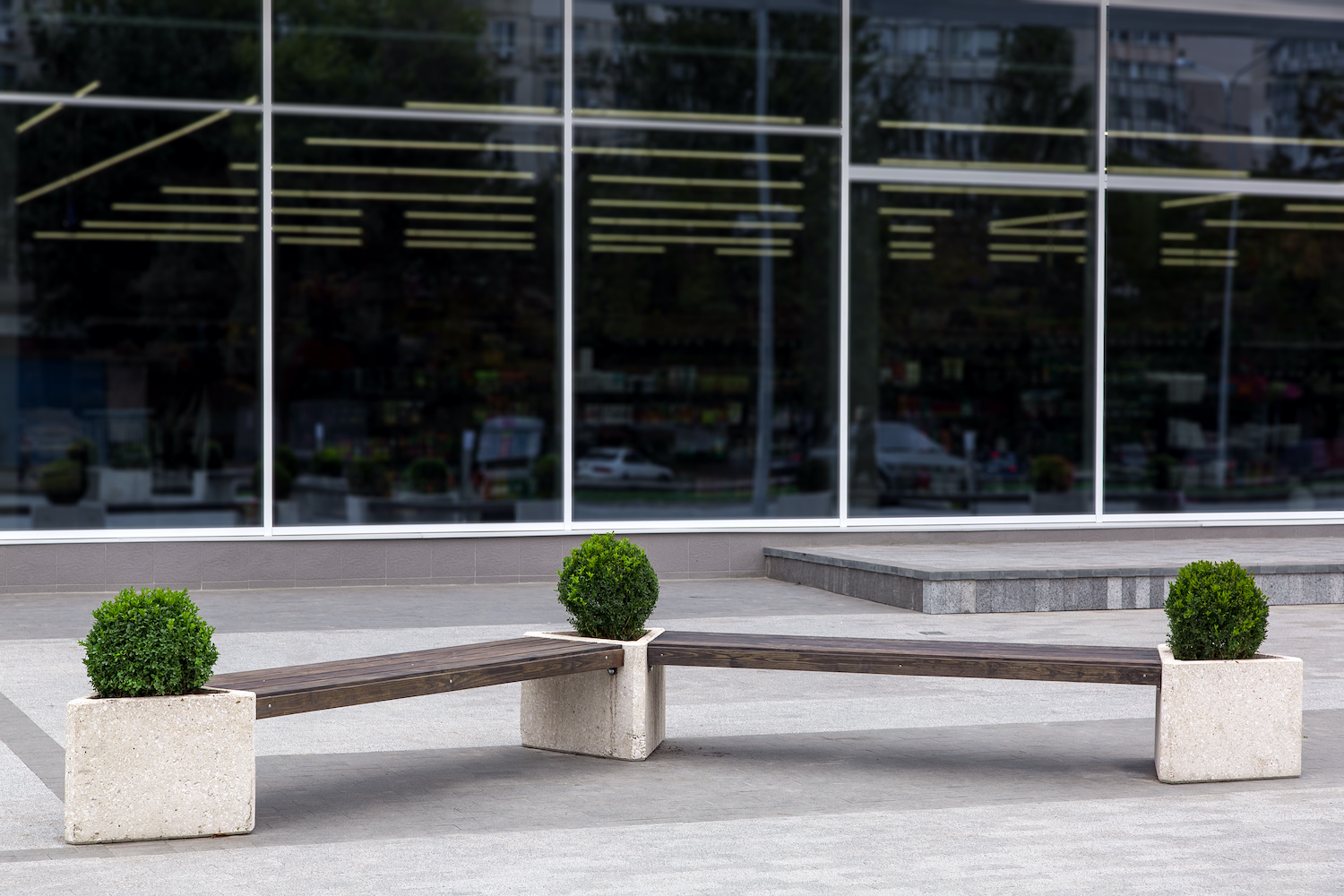 A bench near the facade with panoramic windows of the shopping center, a wooden bench with stone flowerpots for the bushes.